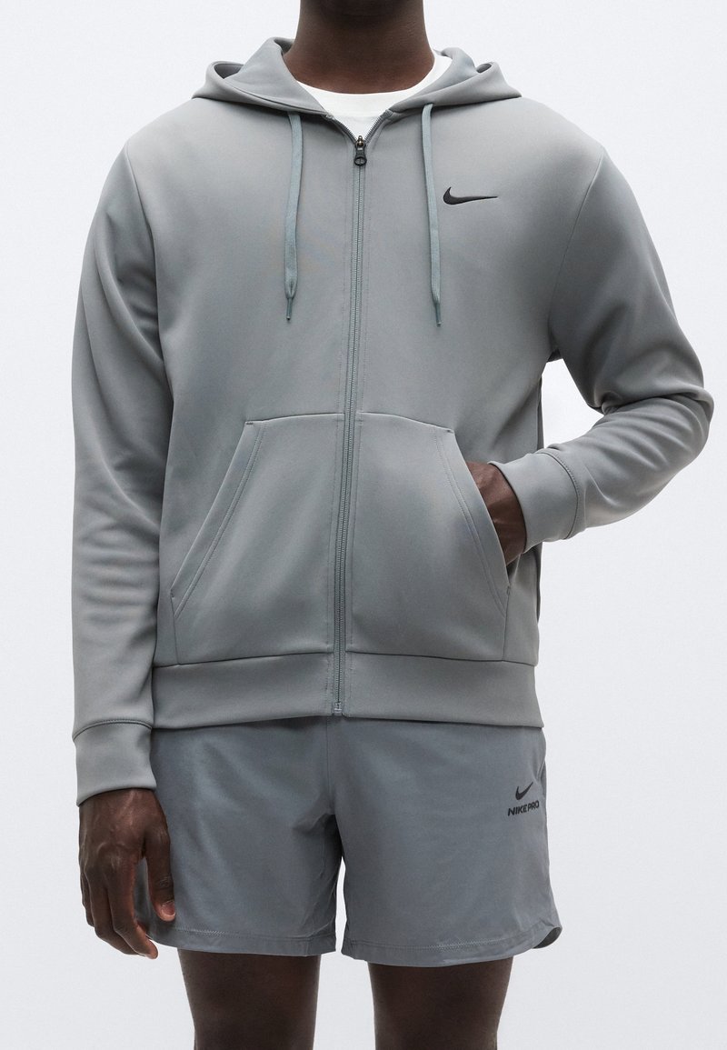 Homme portant un sweat à capuche zippé Nike gris clair et un short assorti, une main dans la poche du sweat, debout sur un fond uni.