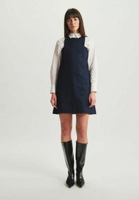 Robe salopette en jean foncé avec un corsage ajusté, jupe à longueur genou et chemise blanche à col. Des bottes noires à hauteur de genou complètent le look.
