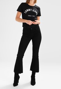Svart t-shirt med vit "TOMMY JEANS" logotyp och texten "SINCE 1985", kombinerad med svarta utsvängda byxor i kordflossa och svarta högklackade stövlar.