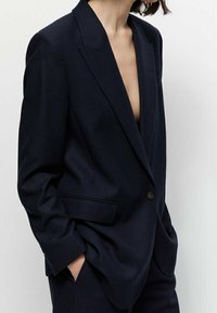 Blazer bleu marine en tissu texturé. Présente un col en V profond, une fermeture à un bouton et deux poches avant. Coupe ajustée.