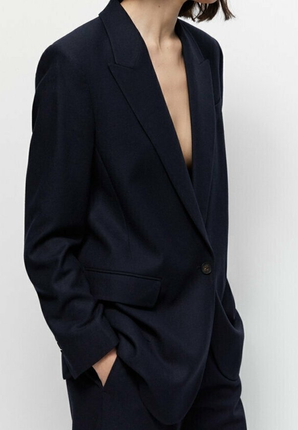 Blazer bleu marine en tissu texturé. Présente un col en V profond, une fermeture à un bouton et deux poches avant. Coupe ajustée.