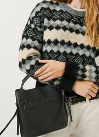 Person der holder en sort lædertaske med præget logo, iført en strikket sweater i sort, grå og beige mønster samt hvide bukser med et sort bælte.