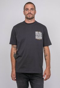 T-shirt en coton gris foncé à manches courtes, col rond, et une poche poitrine avec un design à motifs multiples et un logo en blanc et orange.