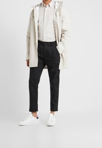 Impermeabile beige chiaro, camicia bianca con colletto, pantaloni neri e sneakers bianche con dettagli neri. Texture liscia con vestibilità casual.