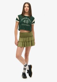 Groene cropped t-shirt met witte accenten en print "SUPERDRY 48 CALIFORNIA," gecombineerd met een gelaagde olijfgroene rok en zwarte schoenen.