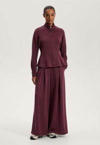 Burgundský loungewear set obsahující zipovou bundu s vysokým límcem a kalhoty s širokými nohavicemi, obojí vyrobené z hladkého, měkkého materiálu.