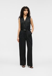 Combinaison sans manches noire à rayures fines avec col, boutonnage à l'avant, taille ceinturée et silhouette à jambes larges. Tissu lisse à rayures verticales.