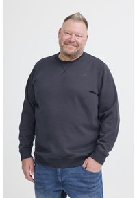 Középkorú férfi, rövid hajjal és szakállal, mosolyogva, sötét színű pulóverben és kék farmernadrágban, az egyik kezét zsebébe dugva, fehér háttér előtt.