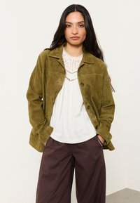 Woman with long dark hair wearing an olive green suede jacket over a white lace-accented blouse and loose dark brown pants, hands in pockets.