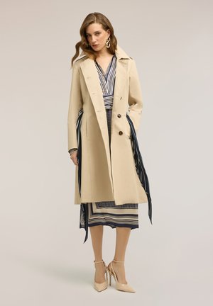 Frau trägt einen beigen Trenchcoat über einem gestreiften marineblauen und weißen Kleid, kombiniert mit beigen hochhackigen Schuhen mit Knöchelriemen, posiert mit einer Hand in der Tasche.