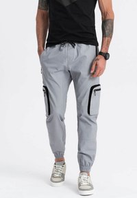 Lichtgrijze cargobroek met elastische boorden, twee zijzakken met zwarte ritsen en een trekkoord in de taille, gedragen met grijze en witte sneakers.