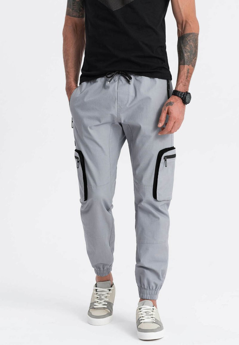 Lichtgrijze cargobroek met elastische boorden, twee zijzakken met zwarte ritsen en een trekkoord in de taille, gedragen met grijze en witte sneakers.