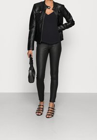 Giacca in pelle nera, top nero a V, pantaloni neri aderenti e sandali con tacco a strappo neri. Tiene una piccola borsa nera.