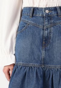 Jupe en denim avec un ourlet froncé. Couleur bleu clair, avec des coutures visibles et un design classique à cinq poches. Comprend une fermeture éclair et un bouton.