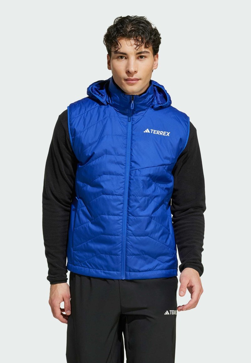 Blå fluffig väst med huva, med ett kviltat mönster och dragkedja fram. Logotyp på bröstet. Bärs över en svart långärmad skjorta.