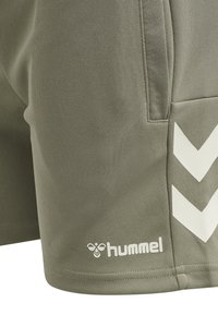 Lähikuva oliivinvihreistä urheilushorteista, joissa on sivutasku, valkoiset viistoviivat ja hummel-logo helman lähellä.