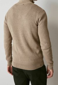 Beige knitted sweater with a high collar, long sleeves, and ribbed cuffs and hem, worn with dark green pants. Smooth texture and fitted shape.