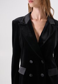 Zwarte fluwelen blazer met satijnen puntrevers, dubbelgeknoopte knopen en zijzakken. Soepele textuur, getailleerde pasvorm en diepe V-hals.