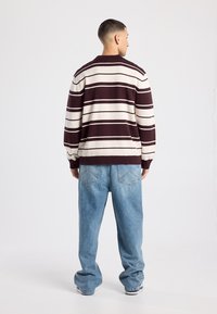 Trui met maroon- en crèmekleurige strepen, voorzien van een geribde kraag en manchetten, gedragen met lichtblauwe losse spijkerbroek, gezien van achteren.