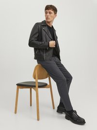 Svart läderbikerjacka med asymmetrisk dragkedja, bärs över en svart skjorta. Grå byxor och svarta loafers, sittande på en trästol.