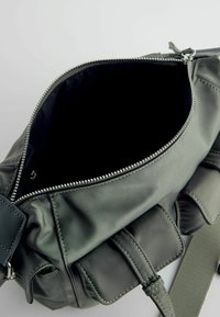 Sac bandoulière vert foncé avec deux poches avant et une fermeture éclair ouverte montrant une doublure intérieure en tissu noir.