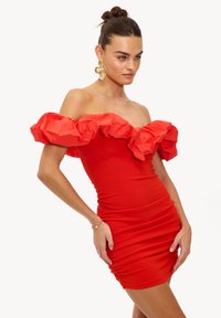 Robe rouge à épaules dénudées avec un col à volants, une coupe ajustée, une texture froncée et une longueur mini. Boucles d'oreilles en or portées.