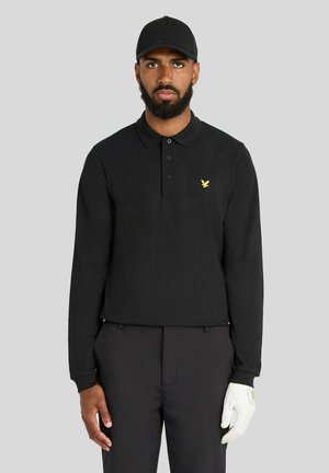 Mann trägt ein schwarzes Langarm-Poloshirt, schwarze Mütze, dunkle Hose und einen weißen Golfhandschuh an der linken Hand vor grauem Hintergrund.