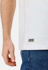 Valkoinen puuvillainen T-paita suoralla leikkauksella, jossa on suorakulmainen logo-patch helman kohdalla. Käsi lepäilee lähellä farkkukangasta vyötäröllä.