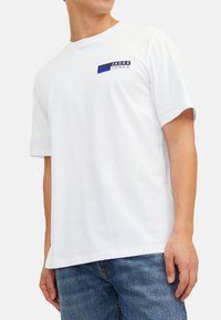Weißes Baumwoll-T-Shirt mit kurzen Ärmeln und rundem Ausschnitt. Mit einem schwarzen und blauen Logo auf der linken Brustseite. Klassische Passform.