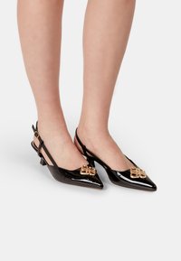 Črne lakirane slingback čevlje s poudarjenim nosom, majhnim bloku peto in z zlatim dekorativnim zaponko na zgornjem delu.