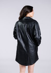 Svart läder skjortjacka med en avslappnad passform, synliga sömmar och rundad nederkant. Har en subtil präglad logotyp på baksidan.