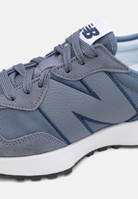 Šedá teniska s kombinací texturované kůže a síťovin. Vybavená šedými tkaničkami a bílou patní částí s logem.