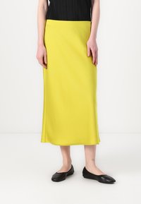 Žlutá midi sukně vyrobená z hladkého materiálu, s vypasovaným pasem a volným střihem, doplněná černými baletními byty. Jednoduchý a elegantní design.