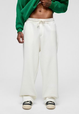 Witte sweatpants met een relaxte pasvorm en een trekkoord in de taille; gecombineerd met een groene hoodie en zwarte sneakers met witte zolen.