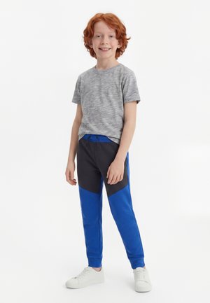 Garçon roux souriant debout portant un t-shirt gris, un pantalon bleu et noir et des baskets blanches sur un fond blanc uni.