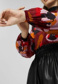 Personne ajustant le poignet d'un chemisier à manches longues aux motifs floraux rouges, roses, orange et noirs, associé à un pantalon taille haute noir.