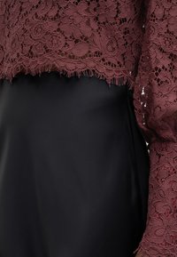 Haut en dentelle bordeaux avec des motifs floraux complexes, doté d'un ourlet à franges, associé à un tissu noir lisse en dessous.