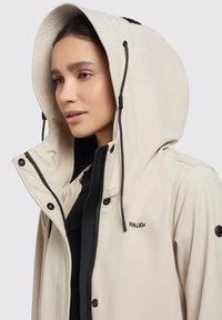 Beige mjukskaljacka med en stor huva, svarta detaljer, framzipper och broderad logotyp. Slät textur och minimalistisk design.
