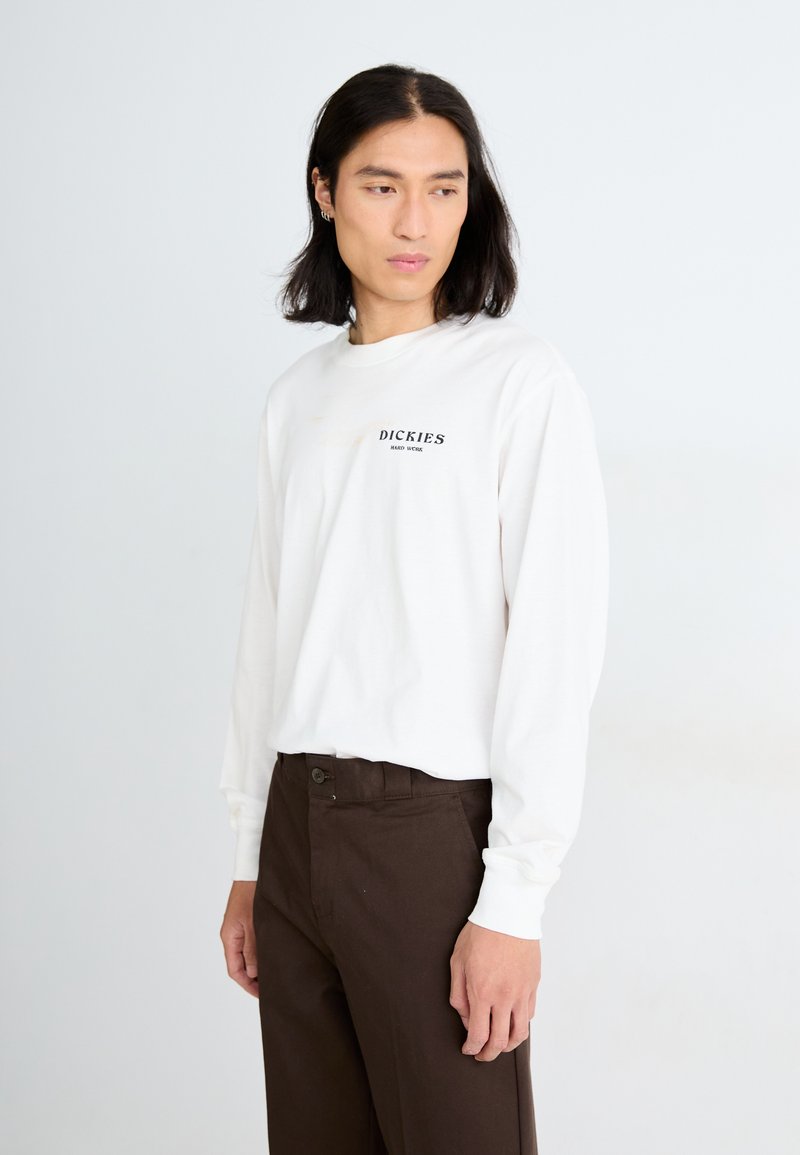 Weißes Langarmhemd mit schwarzem "DICKIES"-Logo auf der Brust, kombiniert mit braunen Hosen. Modell steht leicht seitlich. 