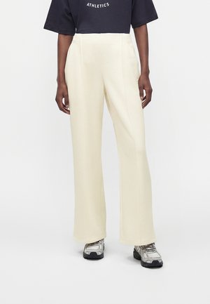 Person iført creme farvede bukser med vide ben, sort kort T-shirt med teksten "ATHLETICS" og grå sportssko står mod en hvid baggrund.