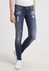 Jean skinny en denim bleu foncé avec des patchs usés et des accents effilochés. Finition avec un design classique à cinq poches et une braguette à boutons.