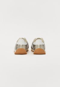 Ténis atléticos prateados e brancos com um traseiro texturizado, sola de borracha gum, e design minimalista, apresentando bordas arredondadas e um perfil baixo.