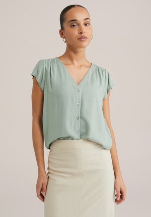 Blusa a maniche corte con scollo a V di colore verde chiaro, con una texture morbida e un motivo sottile; presenta una chiusura con bottoni sul davanti. Abbinata a una gonna color crema.