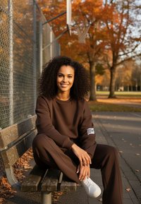 Brązowa bluza i dopasowane spodnie dresowe wykonane z miękkiego materiału, z naszywką z logo. Białe trampki. Park z pomarańczowymi liśćmi.