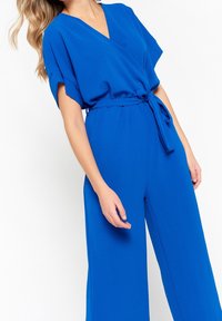 Niebieski kombinezon z głębokim dekoltem w serek, krótkimi rękawami i wiązaniem w talii. Wykonany z gładkiego, lekkiego materiału z szerokimi, zwiewnymi nogawkami.