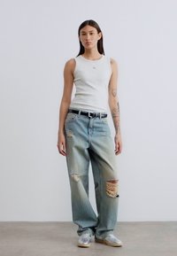 Λευκό αμάνικο τοπ, φθαρμένο ανοιχτό μπλε τζιν με μεγάλες σκισμένες τρύπες, μαύρη ζώνη, γκρι αθλητικά παπούτσια, στέκεται σε ουδέτερο φόντο.