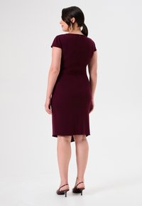 Burgundisches, tailliertes Kleid mit kurzen Ärmeln, knielangem Saum und Reißverschlussdetails hinten; kombiniert mit dunklen, hohen Sandalen. Glatte Stofftextur.