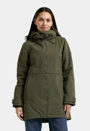 Vrouw draagt een groene winterjas met rits, hoge kraag en trekkoorden, staand tegen een effen lichte achtergrond.