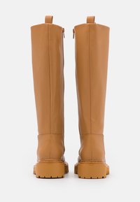 Botas de cuero marrón claro hasta la rodilla con un acabado suave, cremalleras laterales y suelas gruesas y texturizadas. Cuenta con un lazo trasero para facilitar su uso.