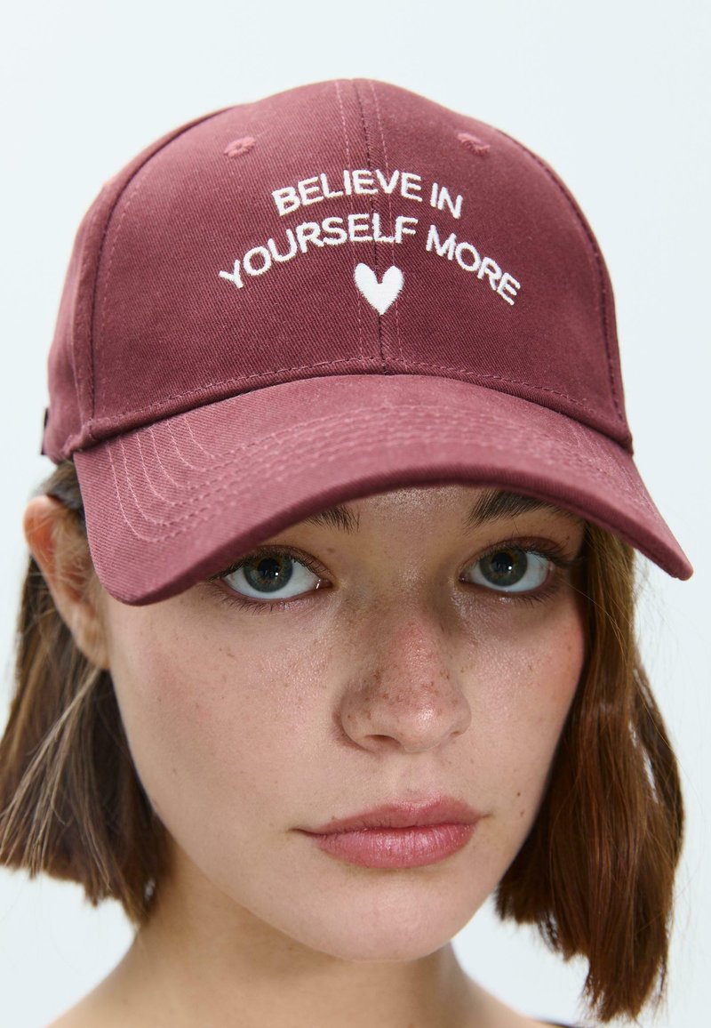 Cappellino da baseball in cotone color bordeaux con testo ricamato bianco "CREDI DI PIÙ IN TE STESSO" e un'icona a forma di cuore. Caratterizzato da una visiera curve.